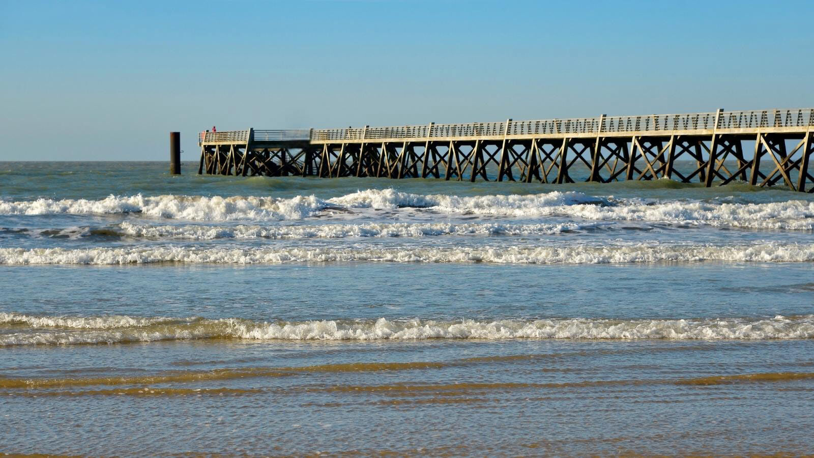 L'estacade de Saint-Jean-de-Monts s'avance sur l'océan Atlantique, avec ses piliers de bois soutenant la promenade, et des vagues s'écrasant sur la plage au premier plan sous un ciel bleu clair.